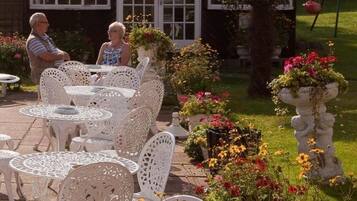Outdoor patio seating with white metal chairs and tables, surrounded by a garden with various flowers and a statue.