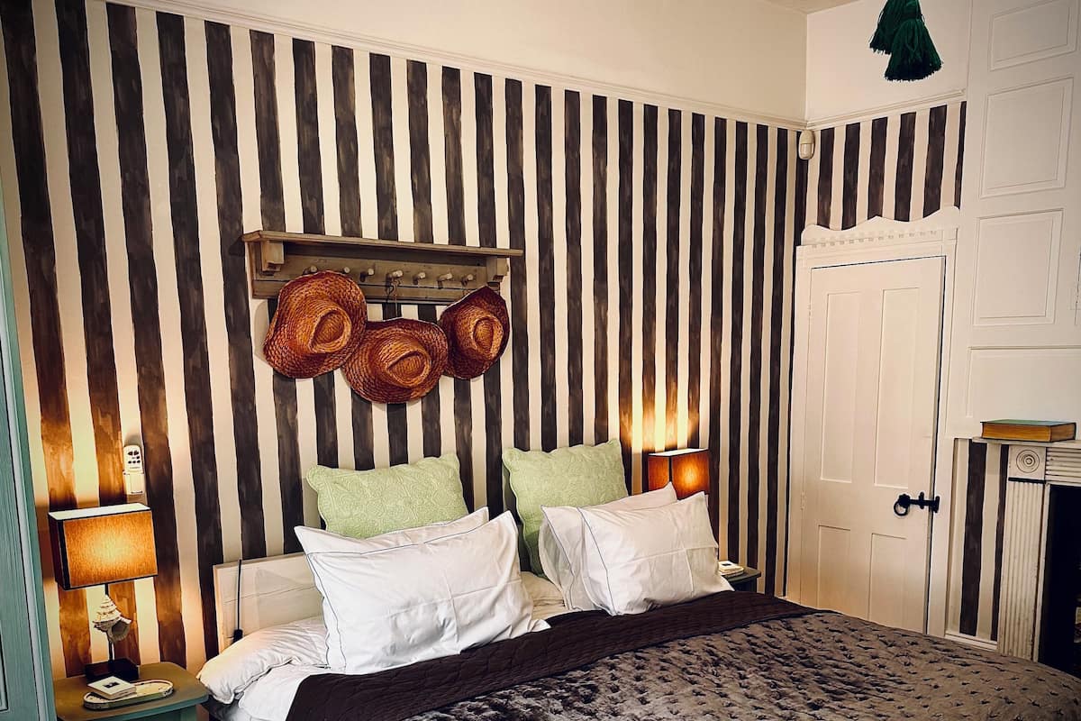 Un dormitorio con papel tapiz a rayas, una cama con almohadas blancas y verdes, un cabecero de madera y una lámpara verde colgante.