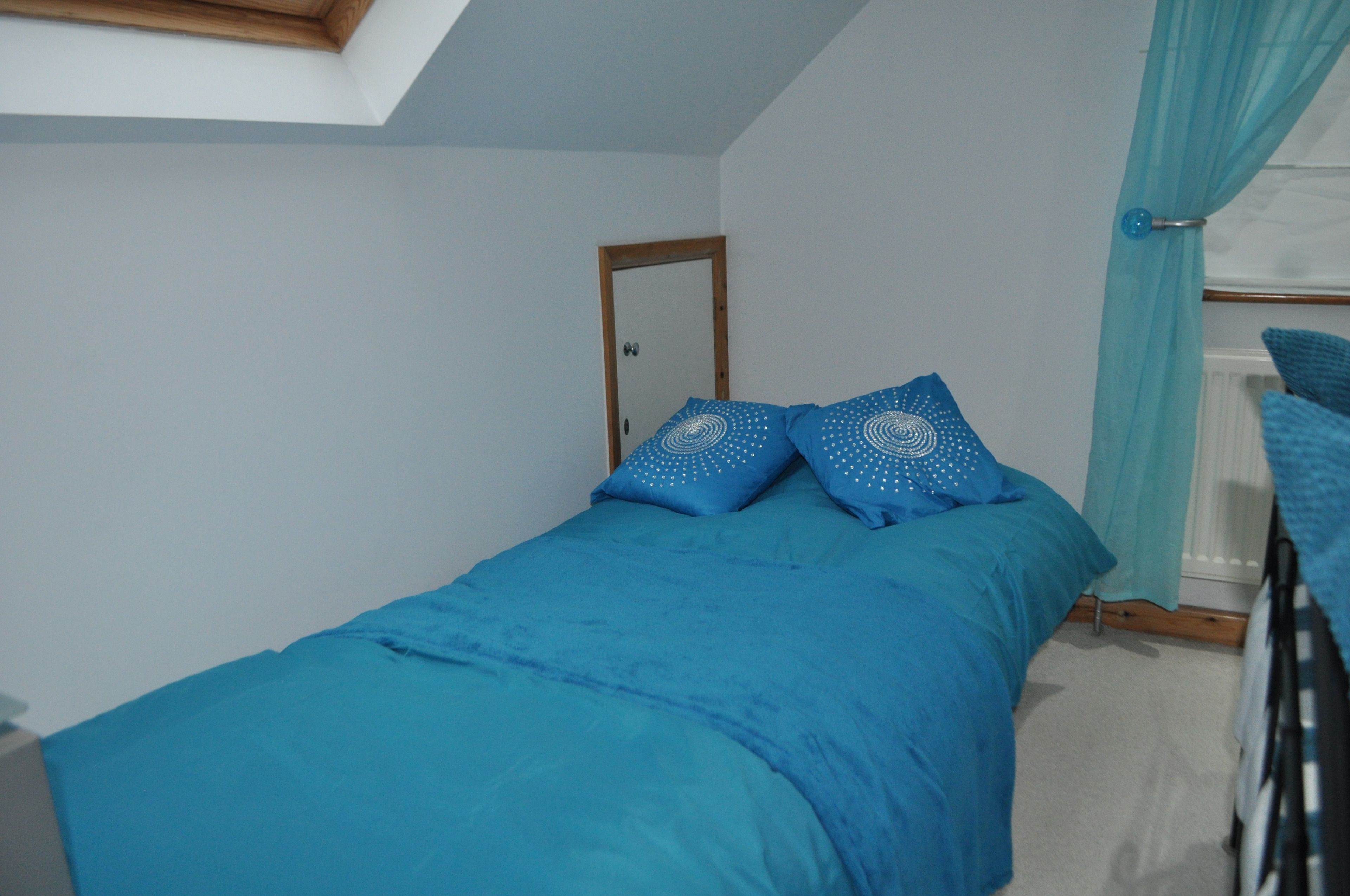 A small, single-bed room with a skylight, a wooden door, and a window with curtains.
