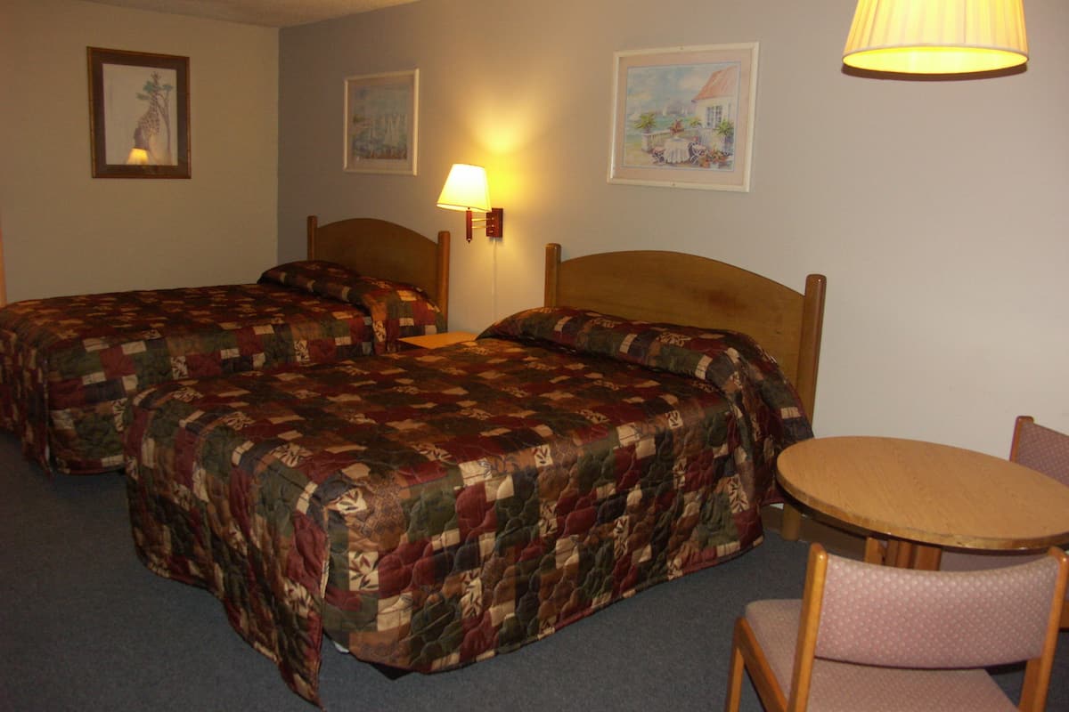 A hotel room with two beds, a round table, and a chair. There are wall-mounted lamps and framed pictures on the walls.