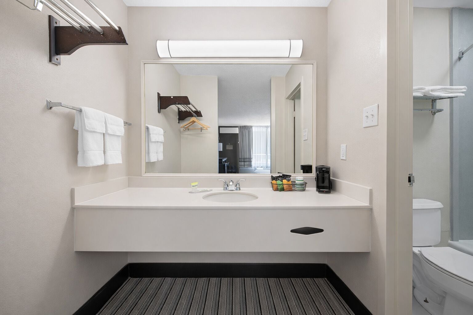Un baño moderno con lavamanos blanco, espejo y toallero. Hay una cafetera y artículos de aseo en la encimera.