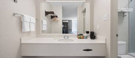 Un baño moderno con lavamanos blanco, espejo y toallero. Hay una cafetera y artículos de aseo en la encimera.