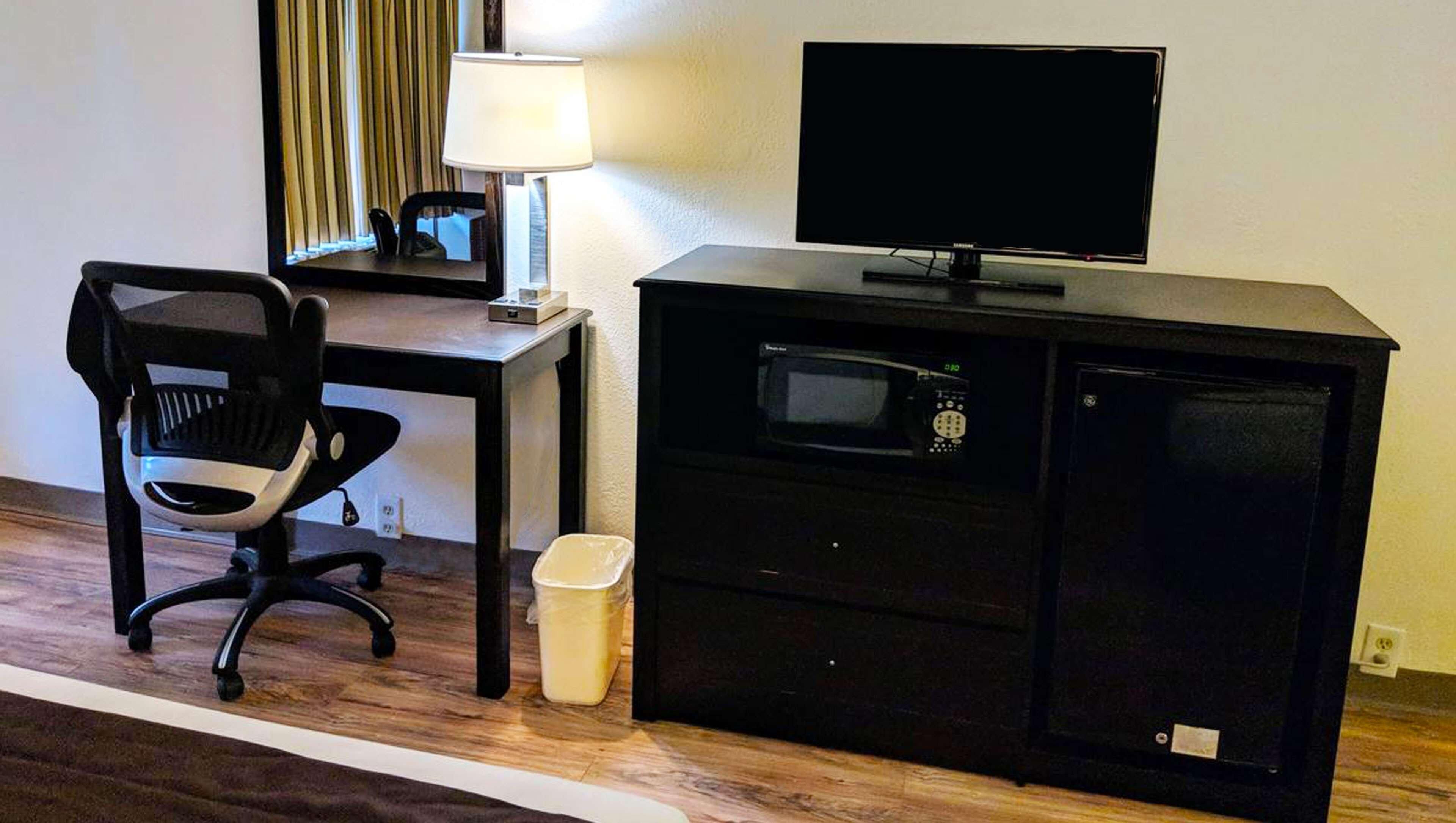 A hotel room with a desk, chair, TV, microwave, and trash bin.