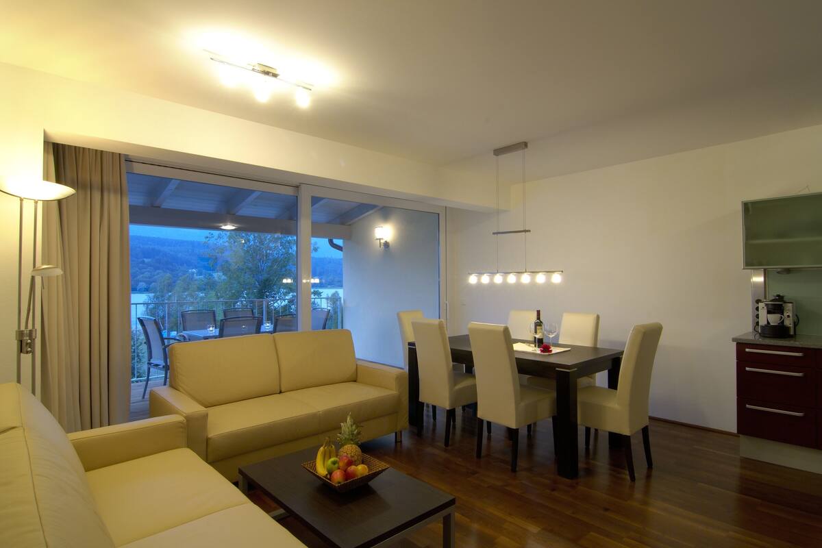 Een moderne woonkamer met een bank, eettafel, stoelen en een schaal fruit. Er is een balkon met uitzicht op bomen en een gebouw.