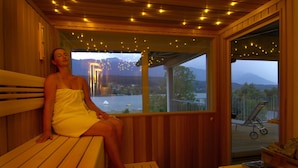 A wooden cabin with a bench and a table, a window with a view of a lake and mountains, and fairy lights on the ceiling. - Appartements De Luxe Schluga (Hermagor-Pressegger See)