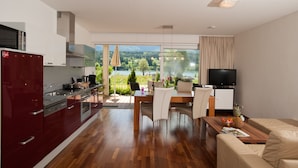 A modern kitchen with a dining area and a living room, featuring a large window with a view of a lake and mountains. - Appartements De Luxe Schluga (Hermagor-Pressegger See)