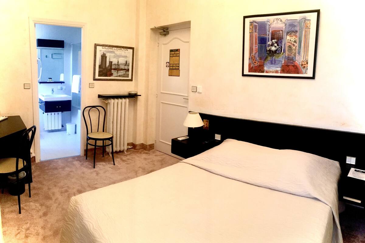 Quarto de hotel com cama, escrivaninha, cadeira, mesa de cabeceira, abajur e um quadro na parede.