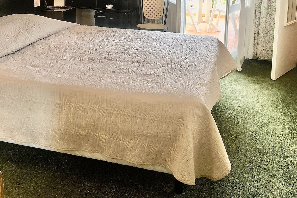 Quarto de hotel com uma cama grande, uma escrivaninha, uma cadeira, um telefone e uma janela com cortinas.