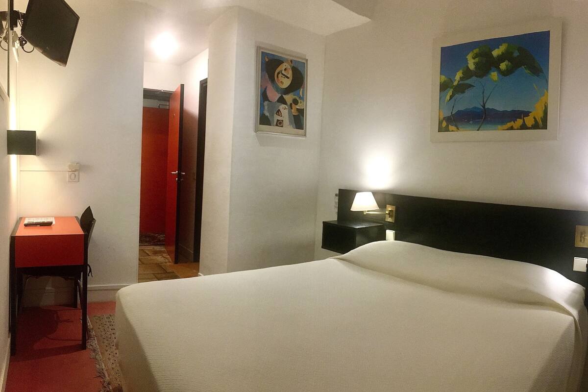 Quarto de hotel com cama, escrivaninha e cadeira, mesa de cabeceira com abajur, e duas pinturas nas paredes.