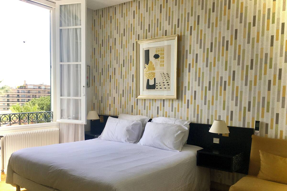 Quarto de hotel com uma cama grande, uma mesa de cabeceira, uma cadeira e uma janela com vista para edifícios e áreas verdes.
