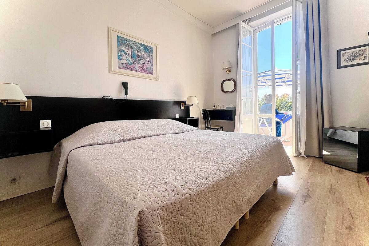 Quarto de hotel com uma cama grande, uma escrivaninha com cadeira, telefone, luminária, quadro na parede e vista para o exterior por uma porta de correr.