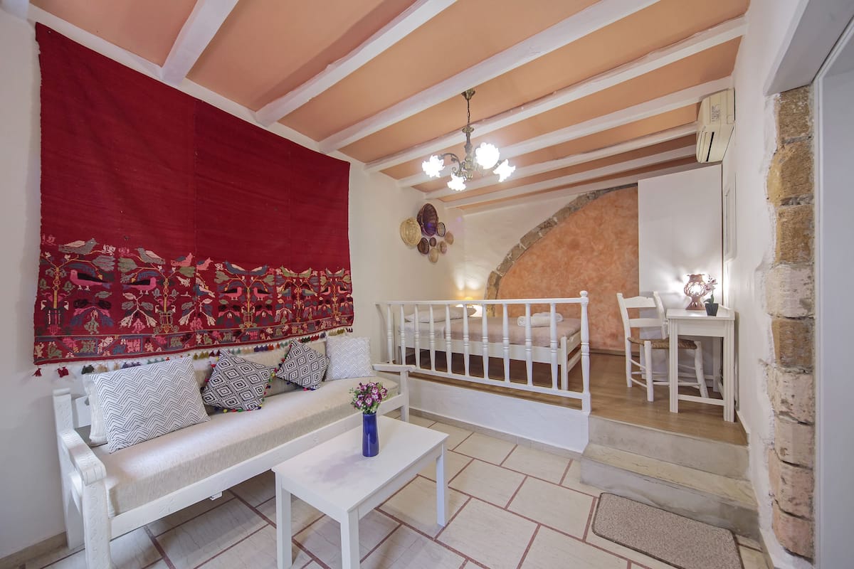 A cozy living area with a white sofa, patterned cushions, a blue vase with flowers, a white coffee table, and a red tapestry.