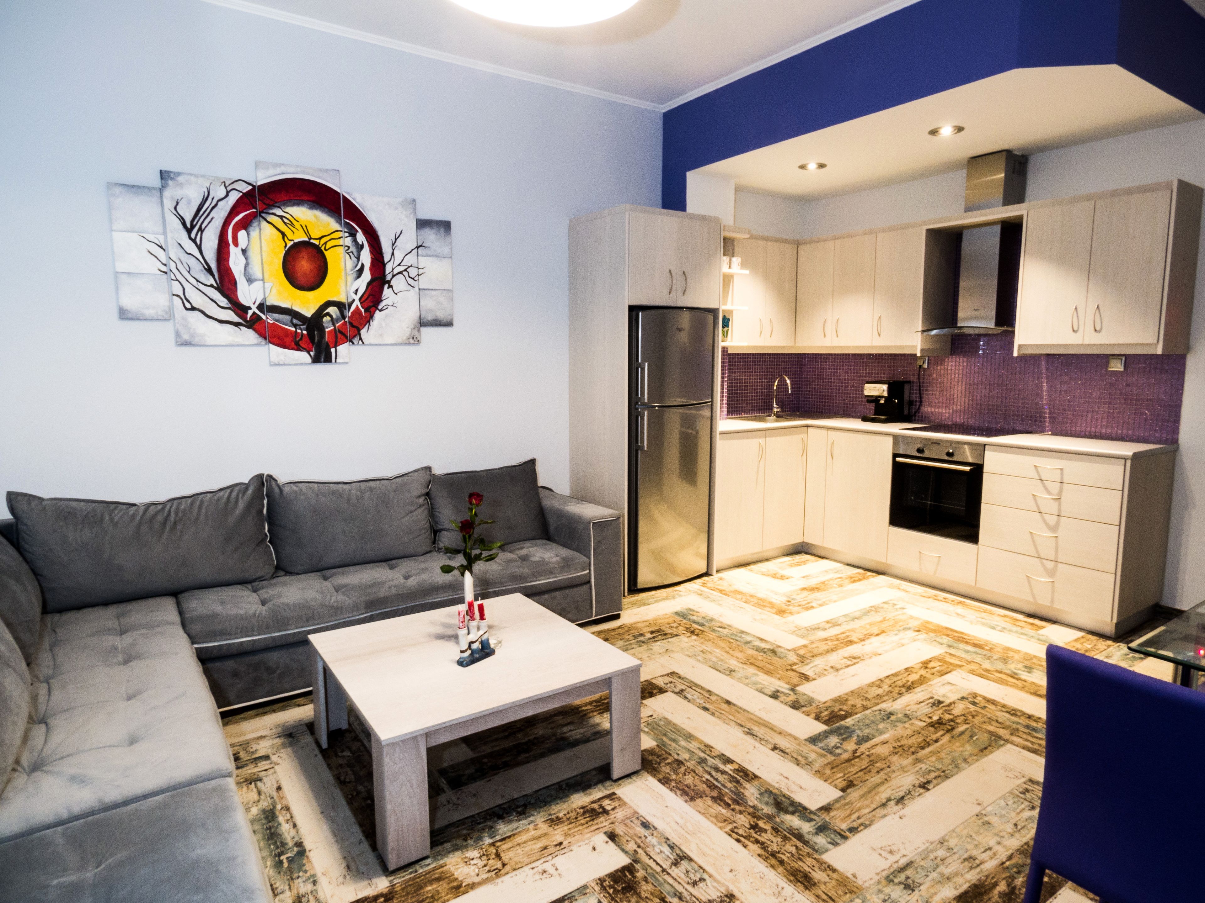 A modern living room with a grey sofa, a coffee table with a vase, and a kitchen with white cabinets and a purple backsplash.