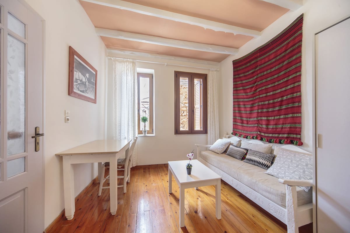A room with a white sofa, a small white table, a wooden floor, a window with curtains, and a red striped curtain.