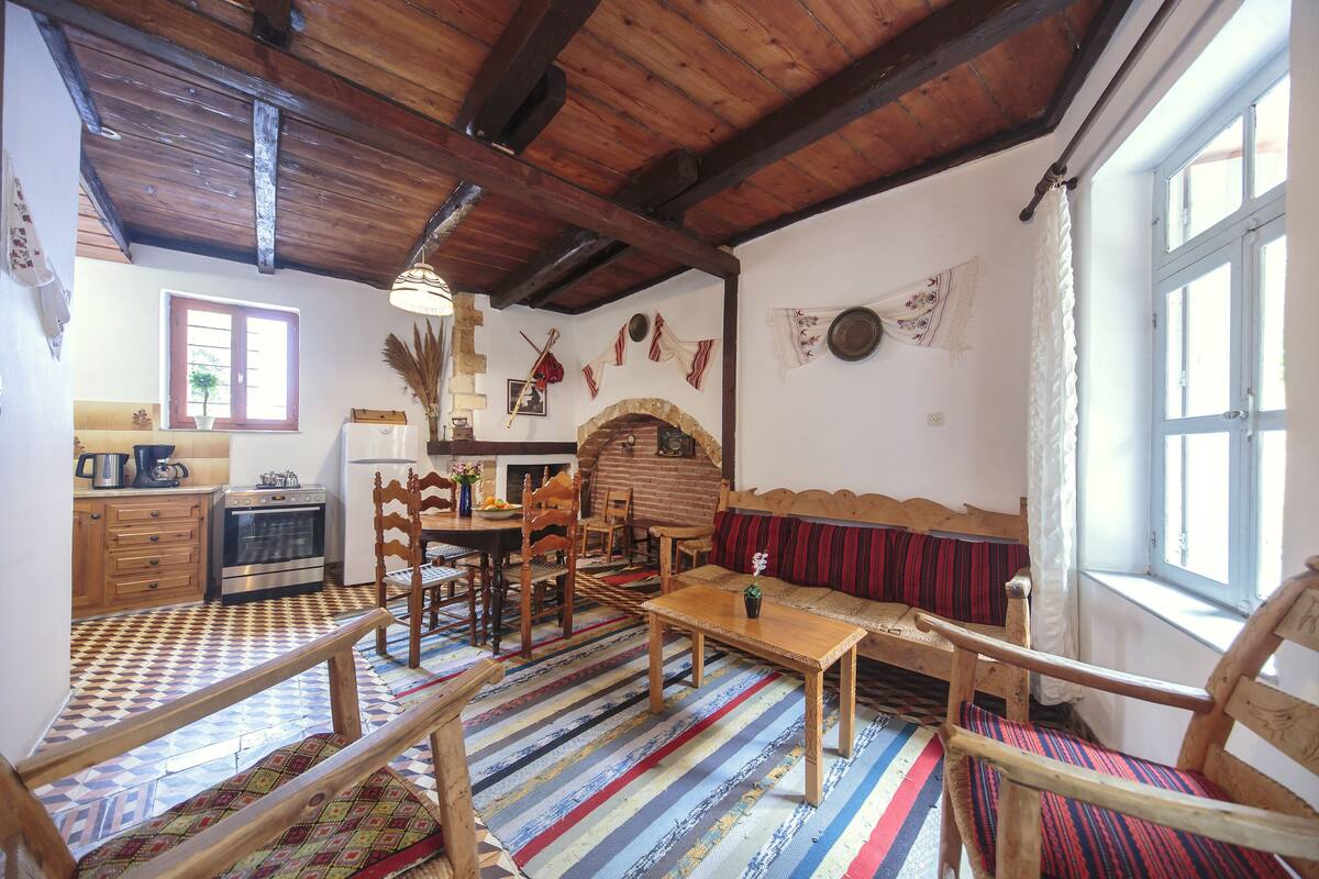 A rustic room with a wooden ceiling, a brick fireplace, a dining area with wooden furniture, and a window with curtains.