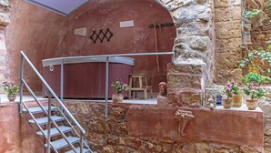 A rustic stone building with a metal staircase leading to a red door. There are potted plants and a small table with a chair outside.