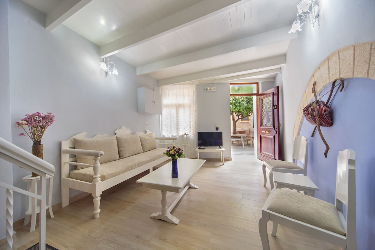 A spacious living room with a sofa, coffee table, and chairs. A TV is mounted on the wall, and there is a door leading to another room.