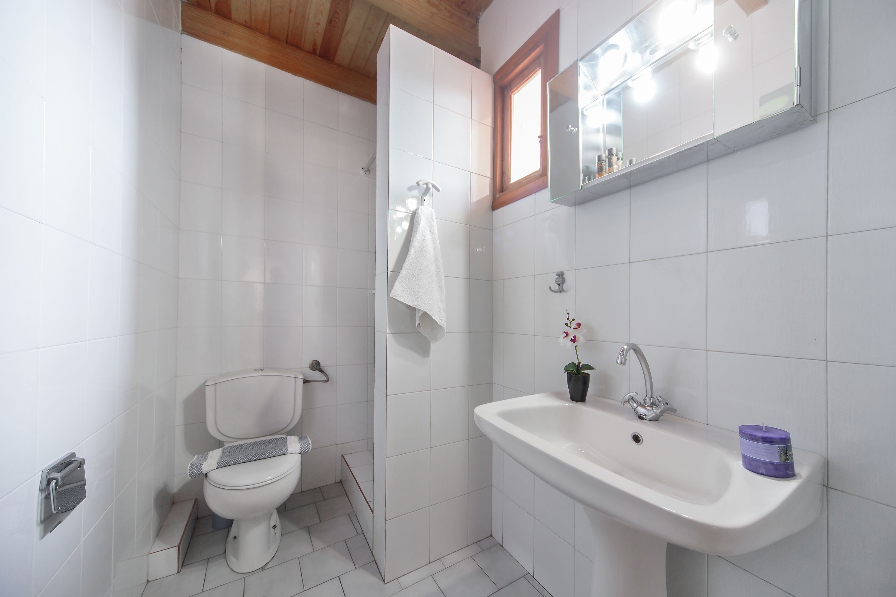 A bathroom with white tiles, a toilet, a sink with a mirror, and a window.