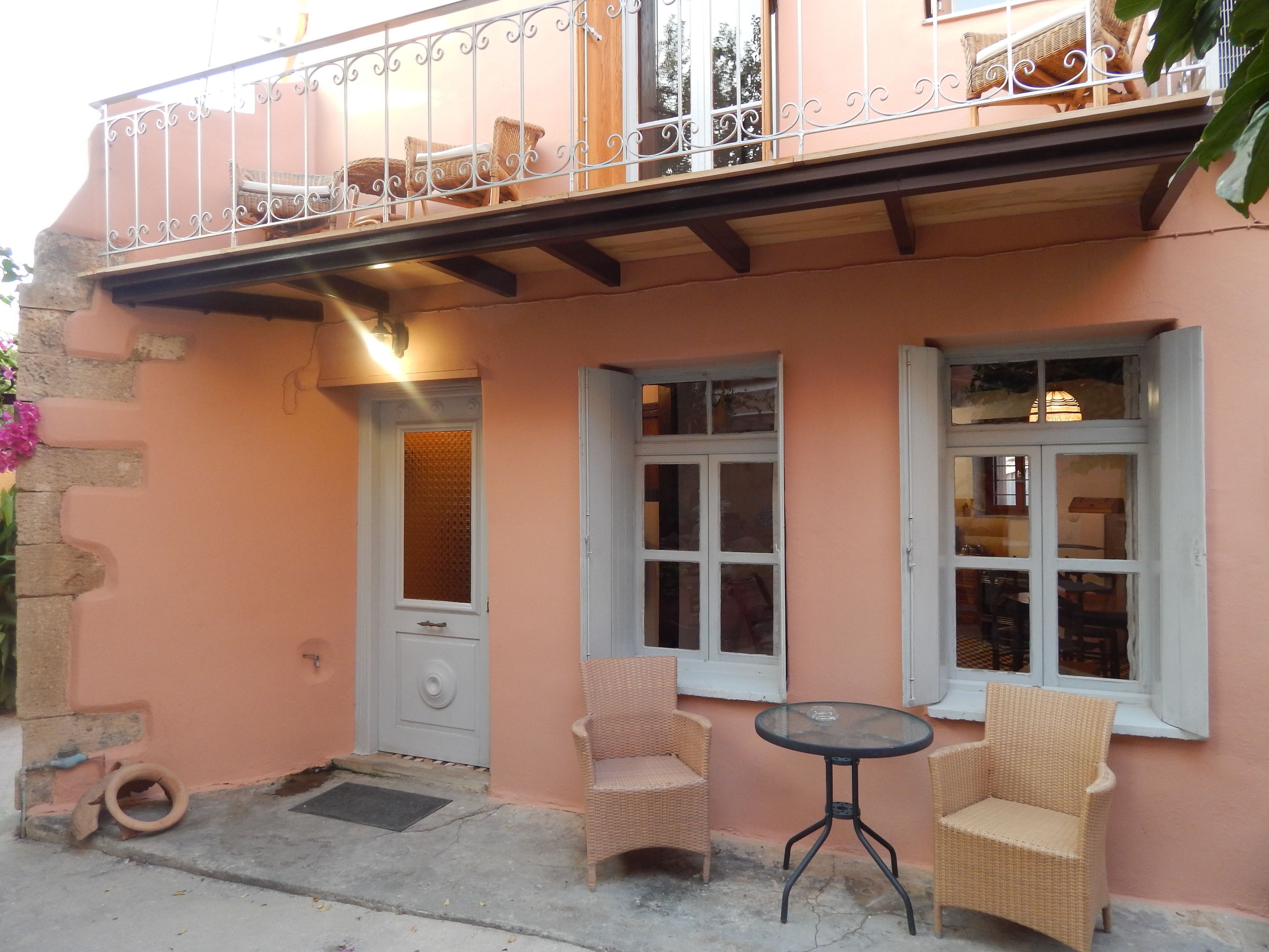 A pink building with a balcony, two wicker chairs, a small round table, and a door with a window.