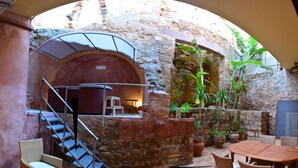 An outdoor patio with a stone wall, a wooden table, wicker chairs, a small staircase, and potted plants.