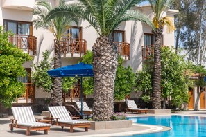 Zona de piscina con sillones reclinables, palmeras y un edificio con balcones.