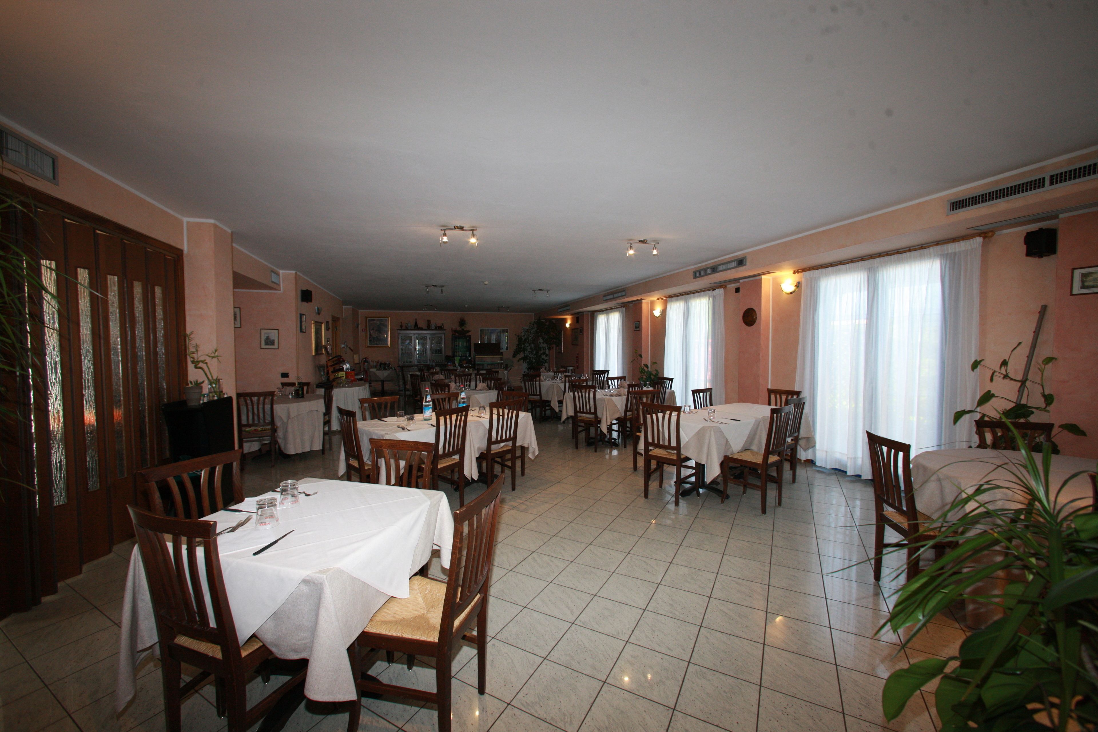 A spacious dining area with multiple tables and chairs, a kitchen in the background, and large windows with curtains.