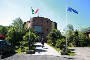 A hotel with a circular brick facade, a flagpole with a flag, and a paved entrance. - Il Canneto (Porto Ceresio)