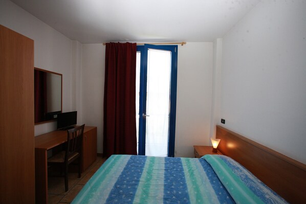 A hotel room with a bed, a desk, a chair, a television, a nightstand with a lamp, and a window with curtains.
