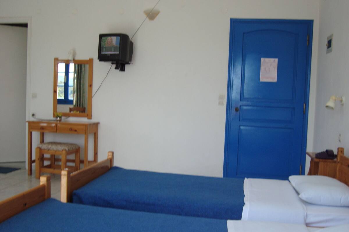 A room with two single beds, a wooden desk with a chair, a television mounted on the wall, and a blue door.