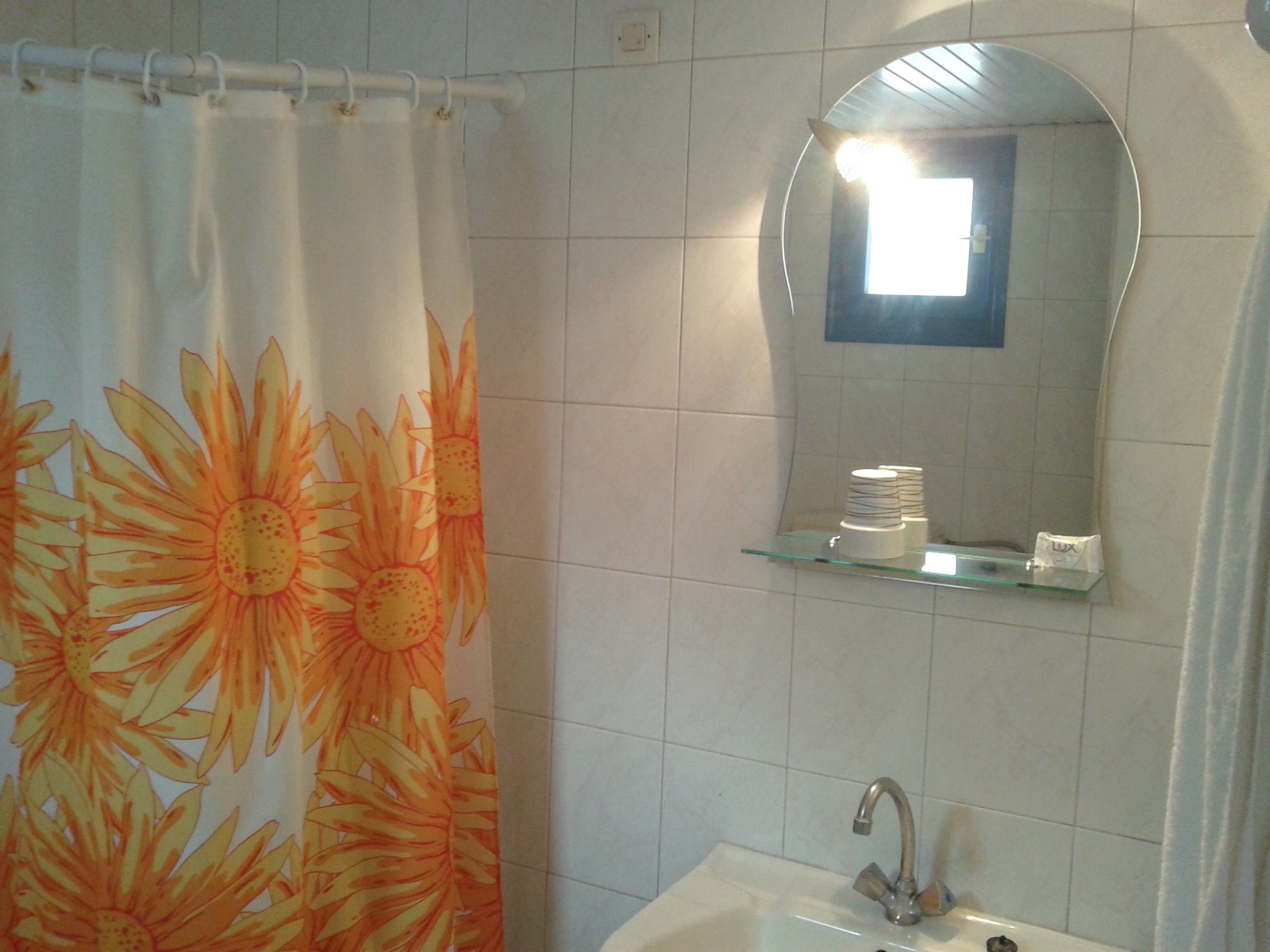 A bathroom with a shower curtain featuring a sun design, a glass shelf above a sink, and a mirror with a light above it.