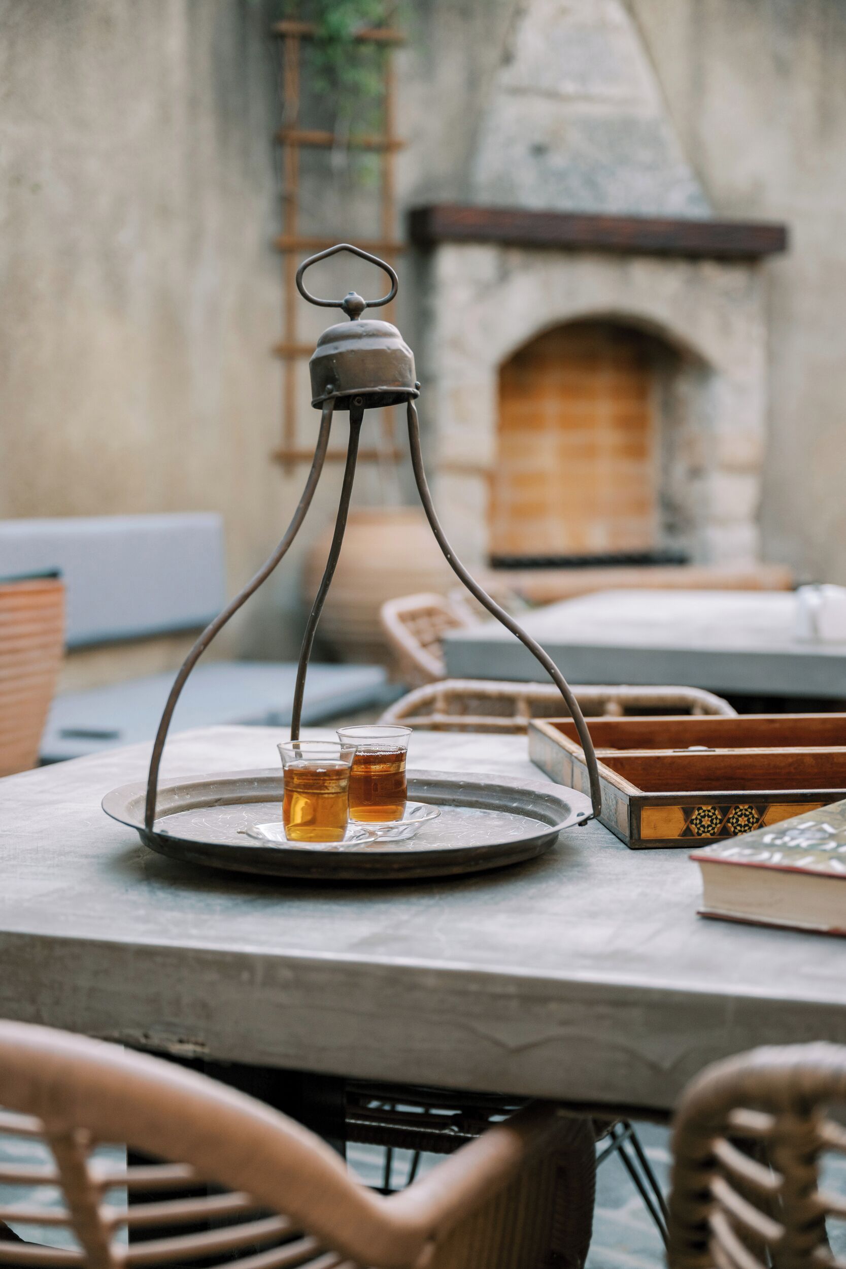 A table with a metal tray holding two glasses of amber liquid, a metal candle holder, and a book. The setting includes wicker chairs and a stone fireplace in the background.