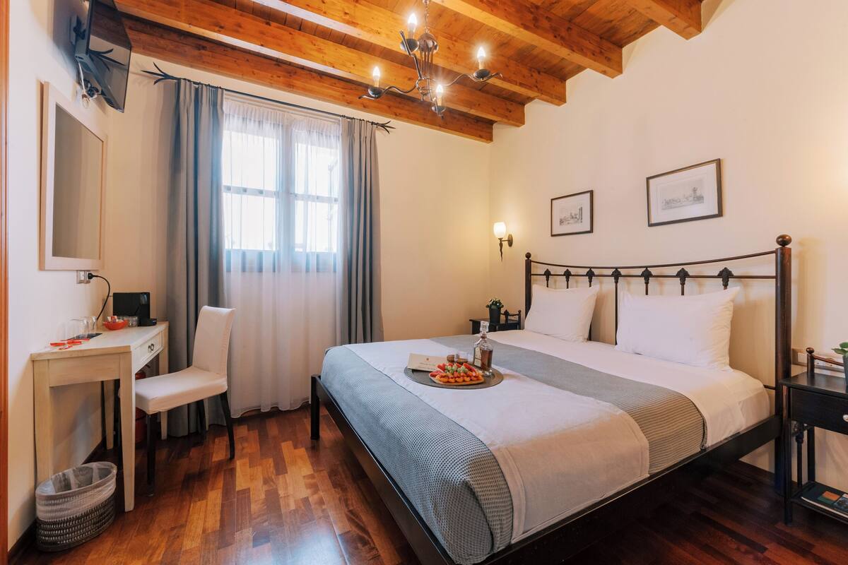 A neatly arranged bedroom with a wooden ceiling, a bed with a tray of fruit, a desk with a chair, and a small table with a plant.