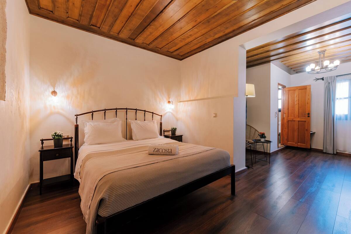 A bedroom with a wooden ceiling, a bed with a 'zacosta' branded pillow, a nightstand with a plant, and a small table with a lamp.