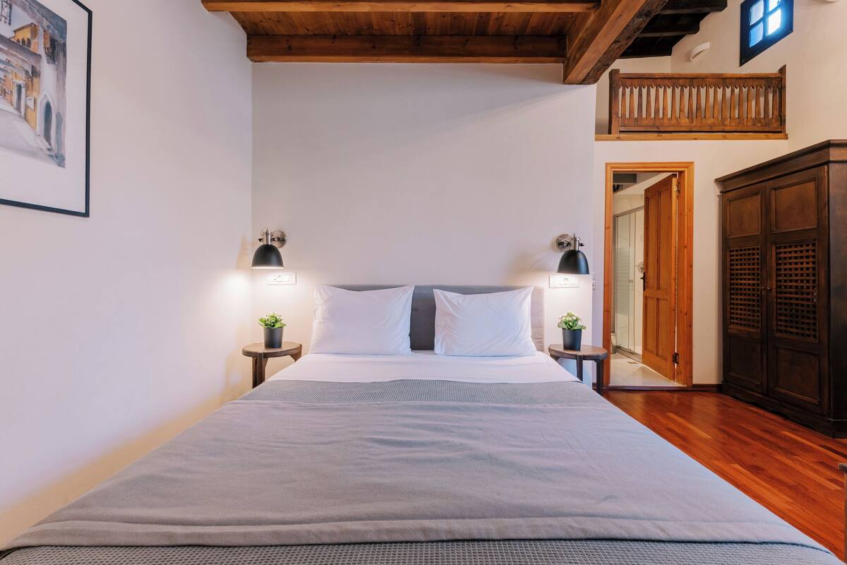 A neatly made bed with a grey blanket, two bedside tables with lamps, a wooden headboard, a wooden wardrobe, and a framed picture on the wall.