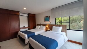 A hotel room with two beds, a nightstand with a lamp, a painting on the wall, and a window with a view of greenery. - Travelers Apartamentos & Suites Castellón de Juanambú (Cali)