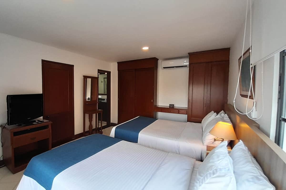 A hotel room with two beds, a TV on a wooden stand, a mirror, and an air conditioning unit.