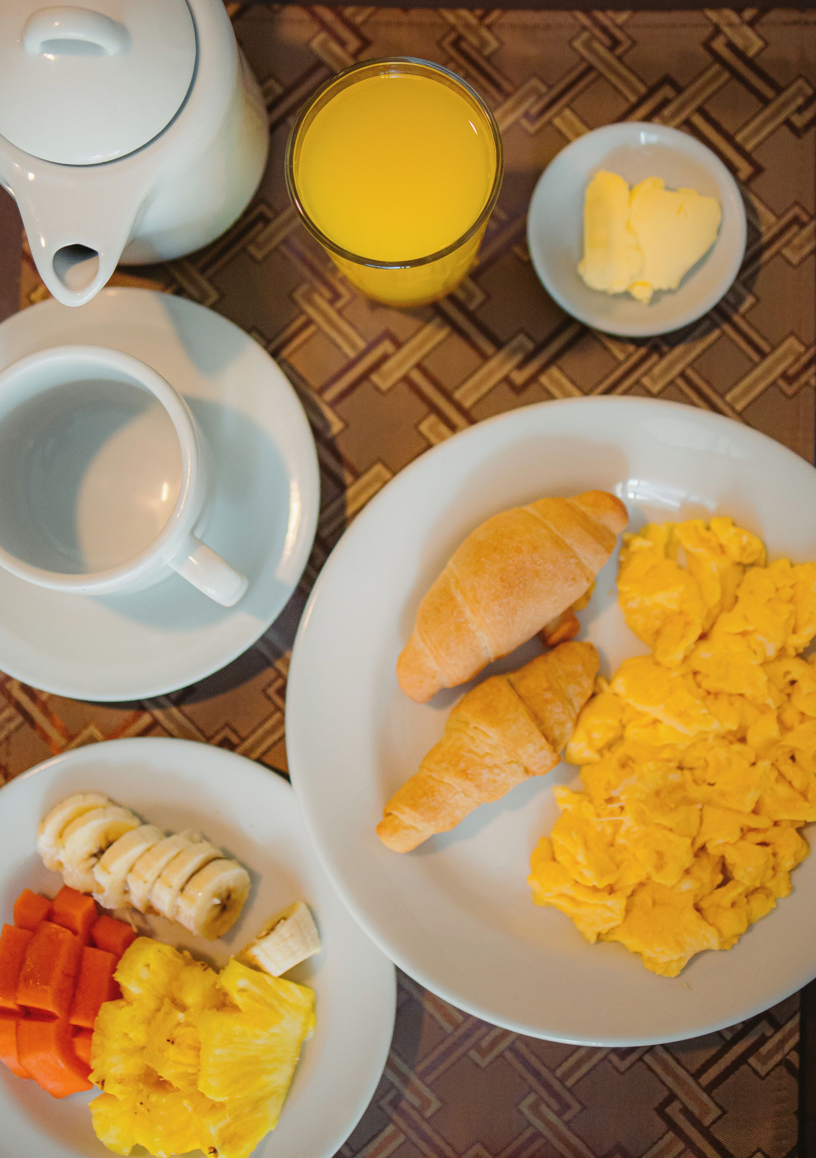 A breakfast table with a teapot, cups, a glass of orange juice, plates of scrambled eggs, croissants, bananas, pineapple, and orange segments.