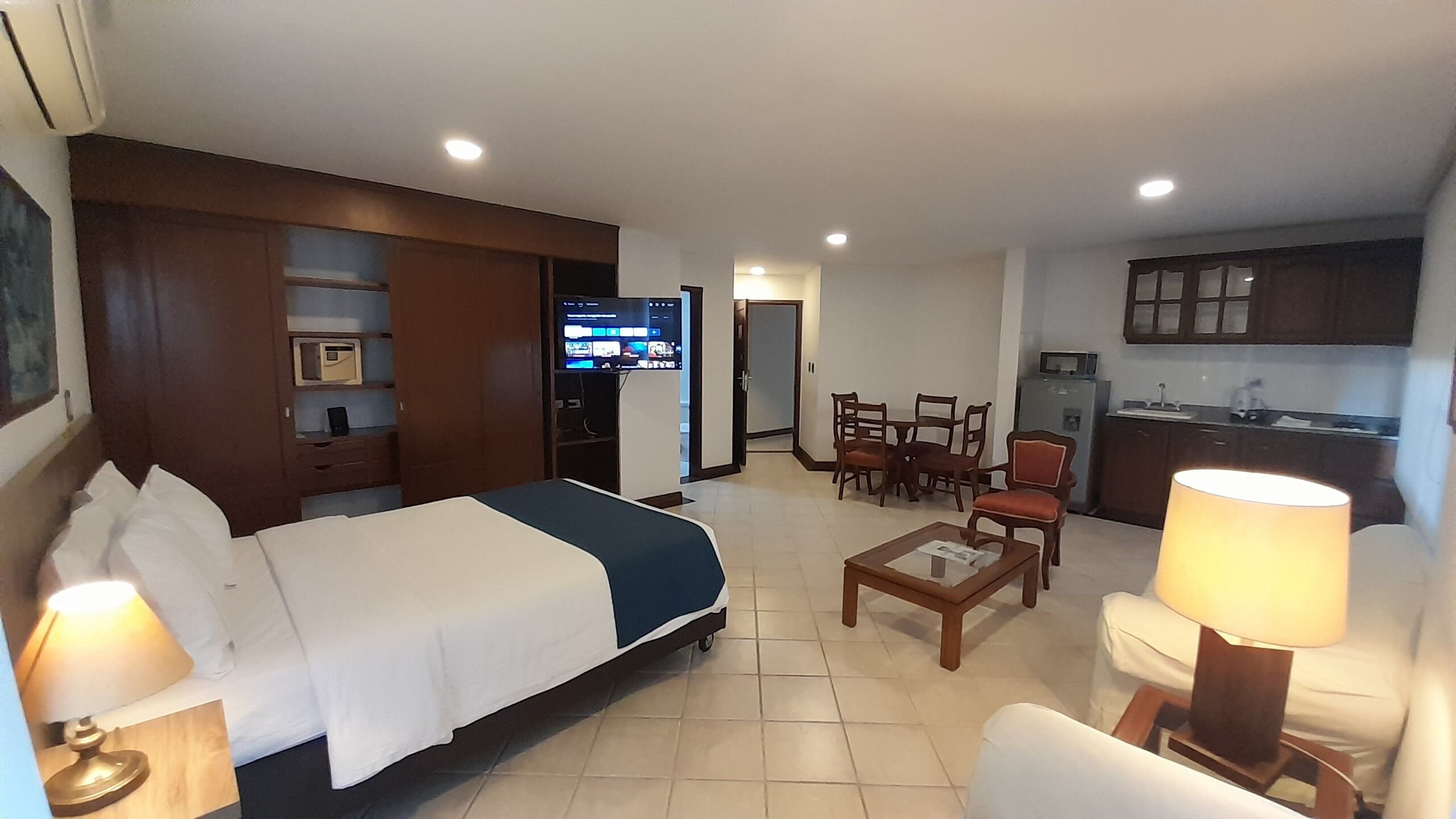A hotel room with a bed, a desk, a chair, a table, a lamp, a TV, and a kitchenette.