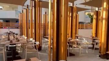 Un área de comedor al aire libre con mesas y sillas, protegida por grandes toldos, rodeada de estructuras de madera verticales.