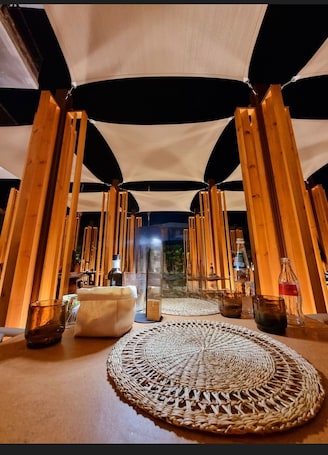 A cozy, tent-like interior with a woven mat at the center, wooden beams, and a glass table with drinks and bottles.