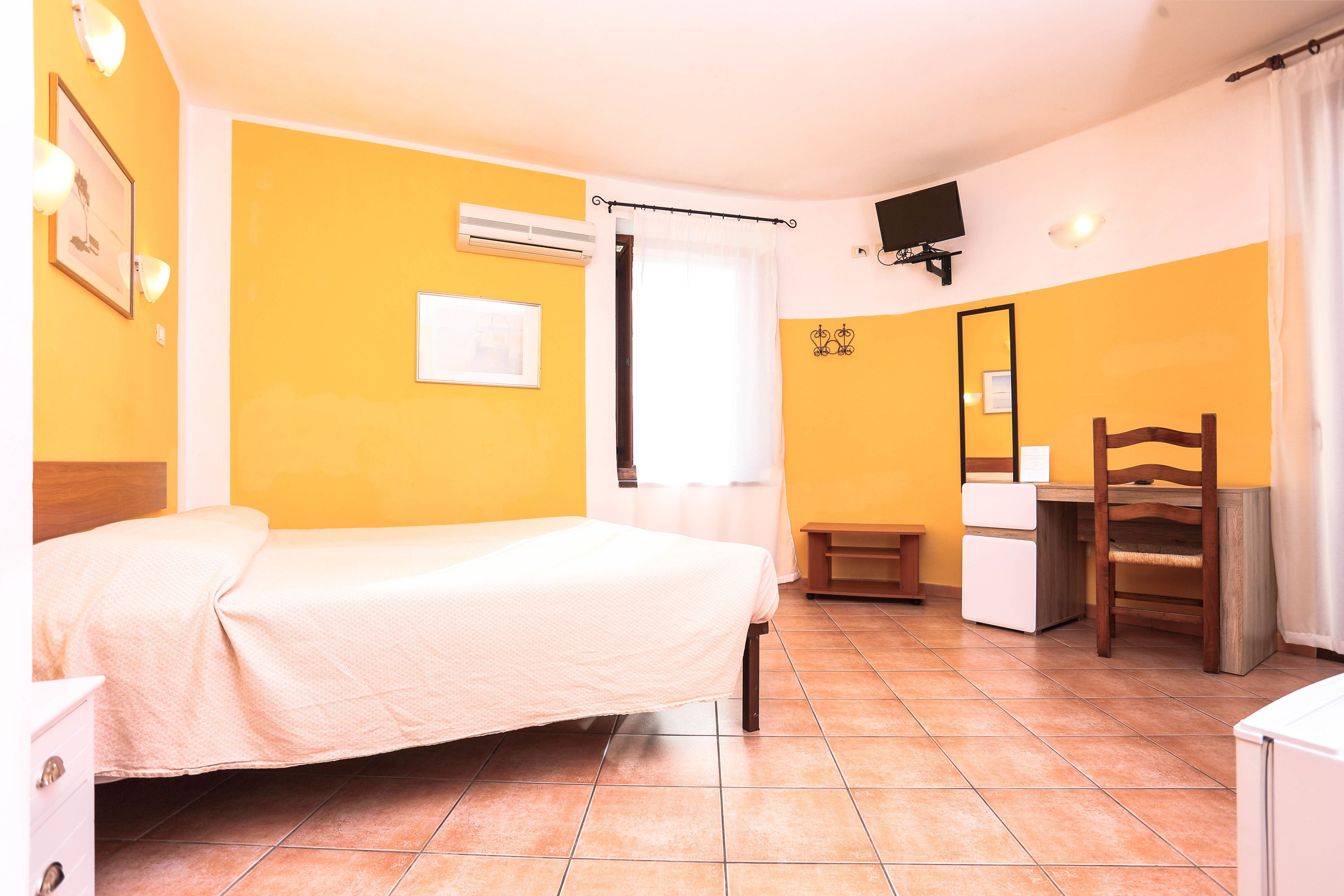Quarto de hotel com uma cama, escrivaninha, cadeira, televisão e espelho.