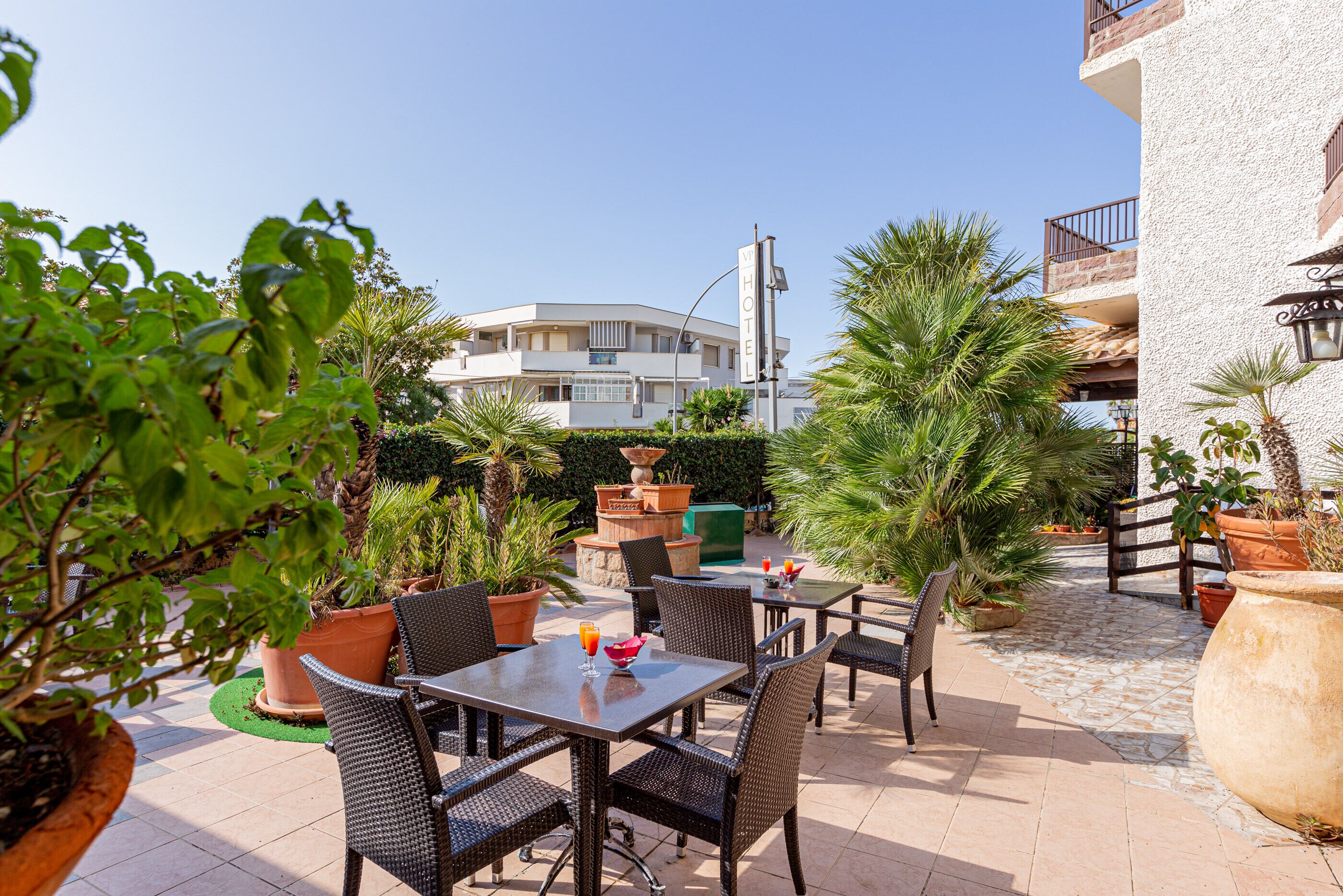 Une terrasse extérieure avec des tables et des chaises en osier, entourée de plantes en pot et offrant une vue dégagée sur un bâtiment d’hôtel en arrière-plan.