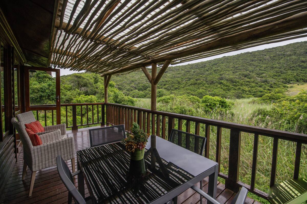 A covered outdoor area with a wooden deck, a wicker chair with a red cushion, a table with a potted plant, and a view of a lush green landscape.