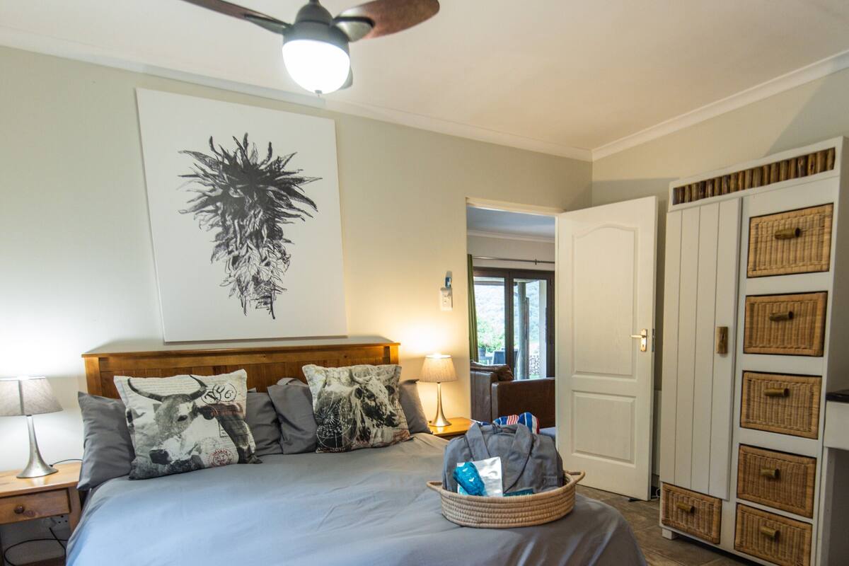 A bedroom with a bed, a ceiling fan, a wall-mounted artwork, a bedside table with a lamp, a nightstand, a wardrobe with baskets, and a door leading to another room.