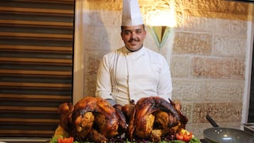 A chef in a white uniform with a tall hat, standing behind a table with two roasted turkeys and a variety of vegetables.