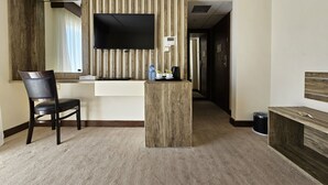 A hotel room with a wooden reception desk, a flat-screen TV mounted on a wooden panel, a chair, and a small table with a water bottle and coffee maker. - Edom Hotel (Wadi Musa)