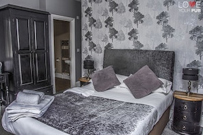 A bedroom with a bed, pillows, a nightstand, a lamp, a wardrobe, and a door.