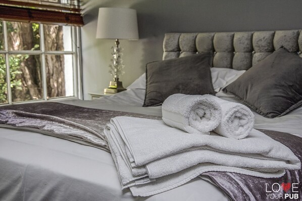 A neatly made bed with rolled towels, a lamp, and a view of trees through a window.
