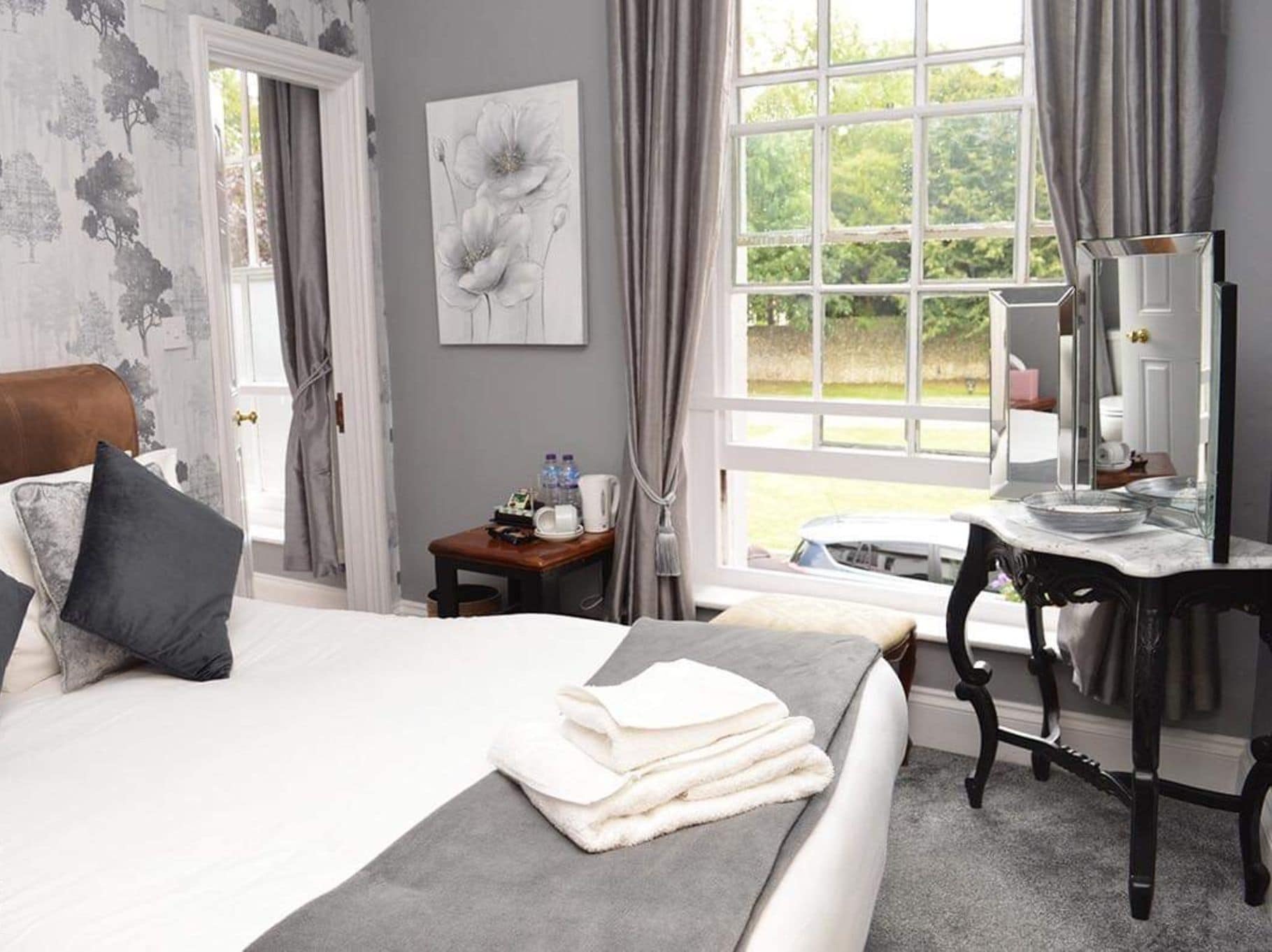 A hotel room with a large bed, a desk, a mirror, and a window with a view of greenery.