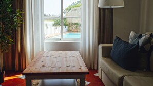 Ein Zimmer mit einem Holztisch, einer Couch mit Kissen, einer Lampe, einem großen Fenster mit Vorhängen und einer Pflanze.